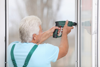 Photo of Mature construction worker repairing plastic window with electric screwdriver indoors