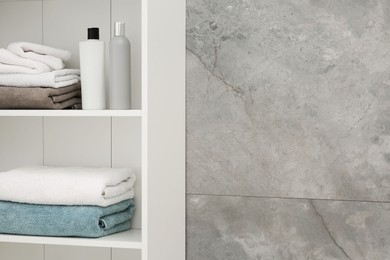 Photo of Cosmetic bottles and terry towels on white rack near light gray marble wall
