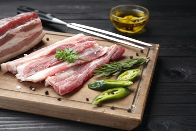 Pieces of raw pork belly, chili pepper, peppercorns, rosemary and parsley on black wooden table, closeup