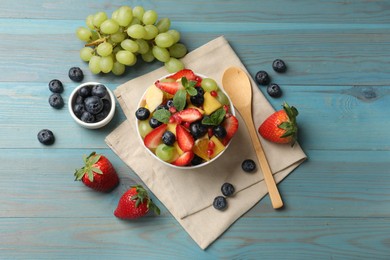 Tasty fruit salad in bowl, ingredients and spoon on light blue wooden table, flat lay