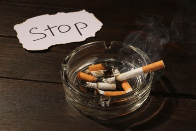 Photo of Ashtray with burnt cigarettes and word Stop written on paper on wooden table, closeup. No smoking concept
