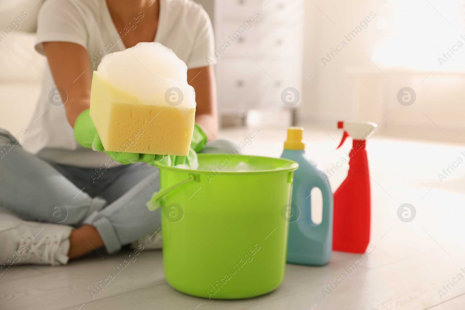 Photo of Woman holding sponge with foam over bucket indoors, closeup. Cleaning supplies