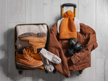 Photo of Open suitcase with warm clothes and camera on wooden floor, flat lay. Winter vacation