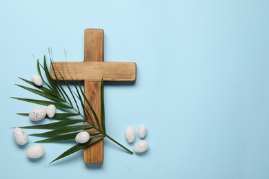 Wooden cross, painted Easter eggs and palm leaf on light blue background, flat lay. Space for text