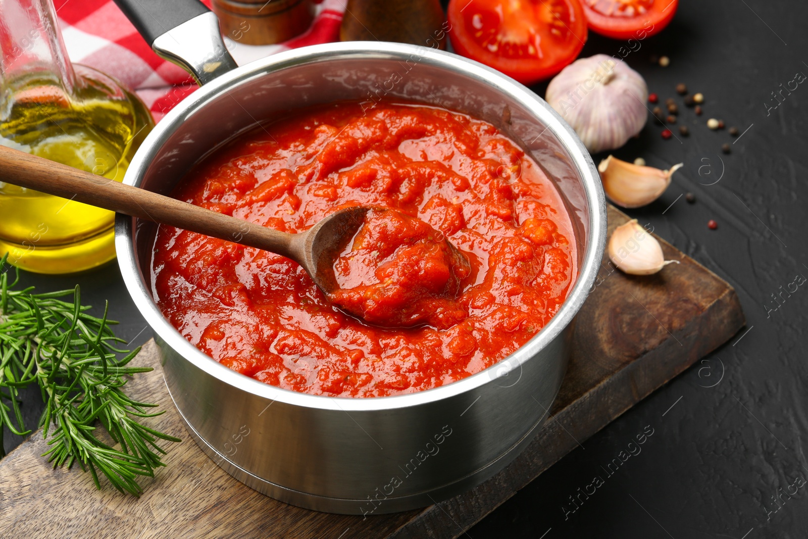 Photo of Homemade tomato sauce in pot, spoon and ingredients on dark table