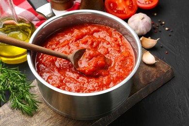 Photo of Homemade tomato sauce in pot, spoon and ingredients on dark table