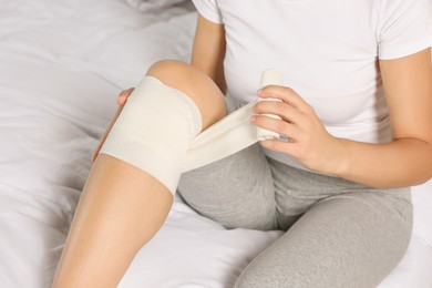 Photo of Young woman applying medical bandage onto knee on bed, closeup