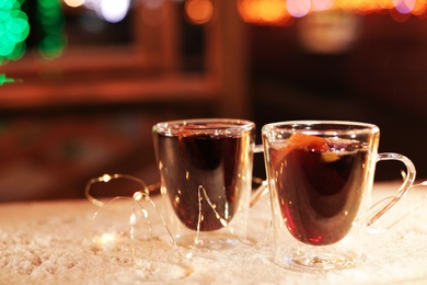 Glass cups of mulled wine and garland on table covered with snow outdoors. Space for text