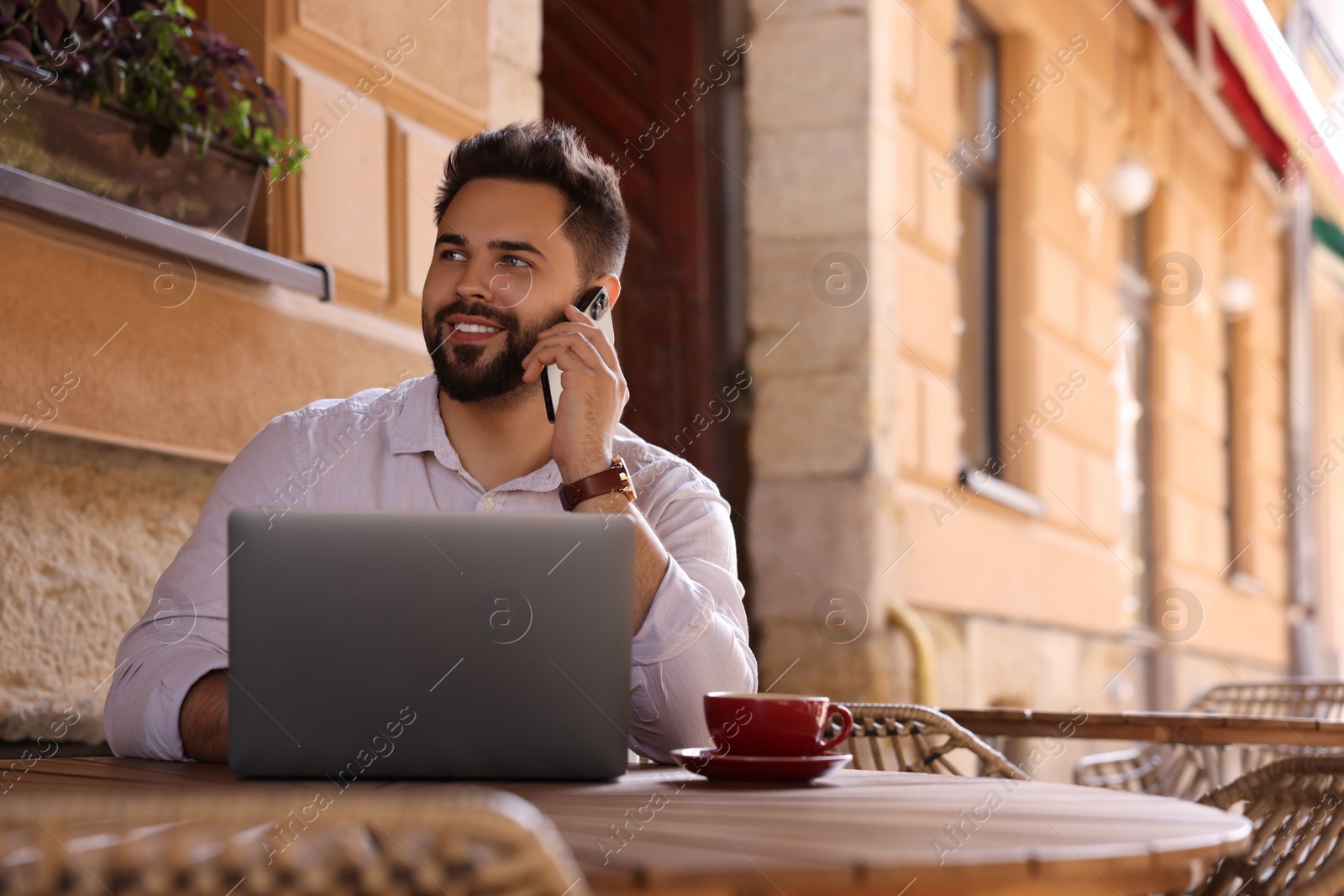 Photo of Handsome young man talking on smartphone while using laptop at table in outdoor cafe. Space for text