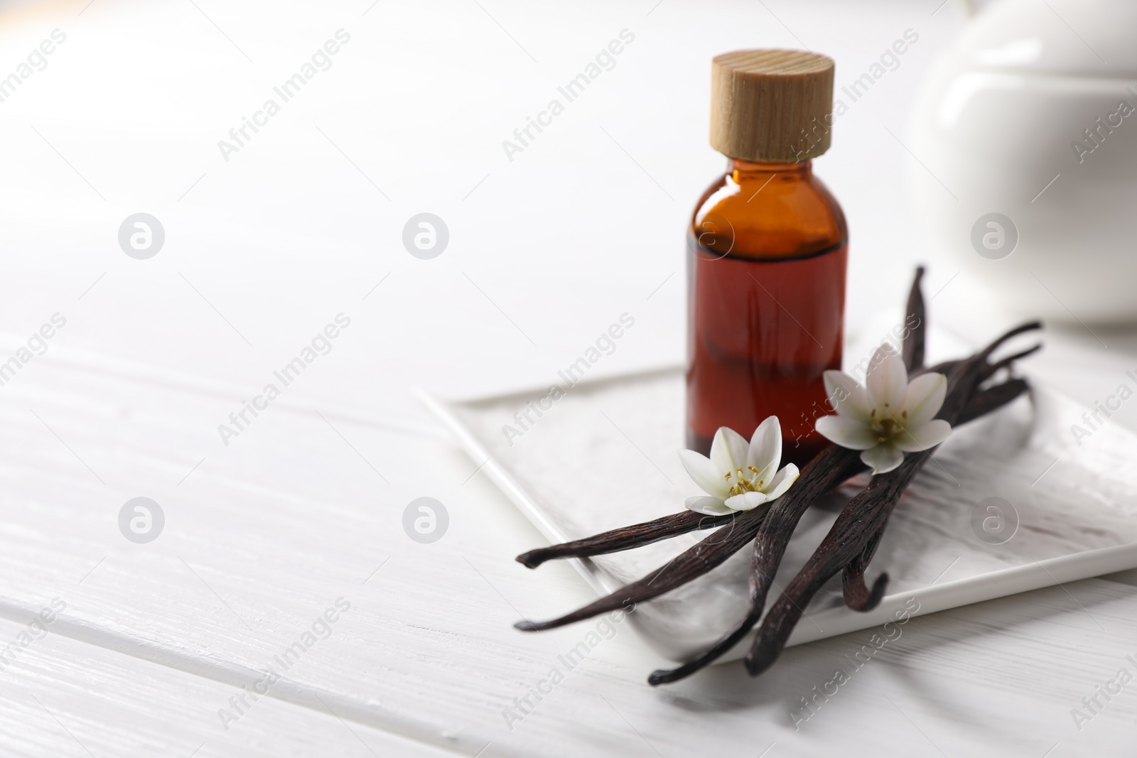 Photo of Vanilla pods, essential oil and flowers on white wooden table, space for text