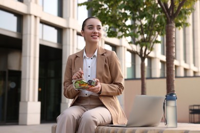 Photo of Happy businesswoman with container of salad having lunch on bench outdoors