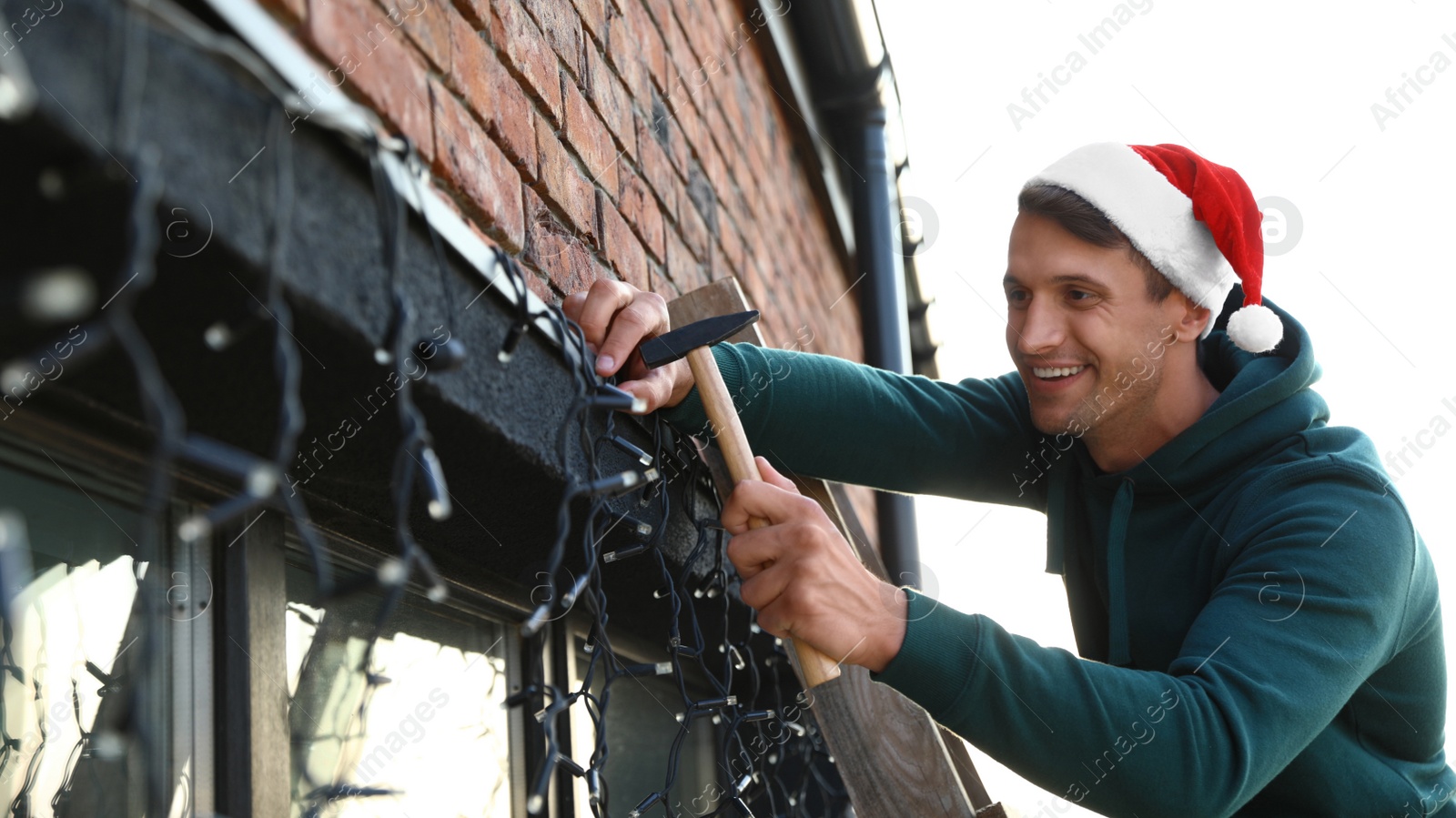 Photo of Man in Santa hat decorating house with Christmas lights outdoors