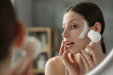 Photo of Young woman washing face with brush and cleansing foam near mirror in bathroom