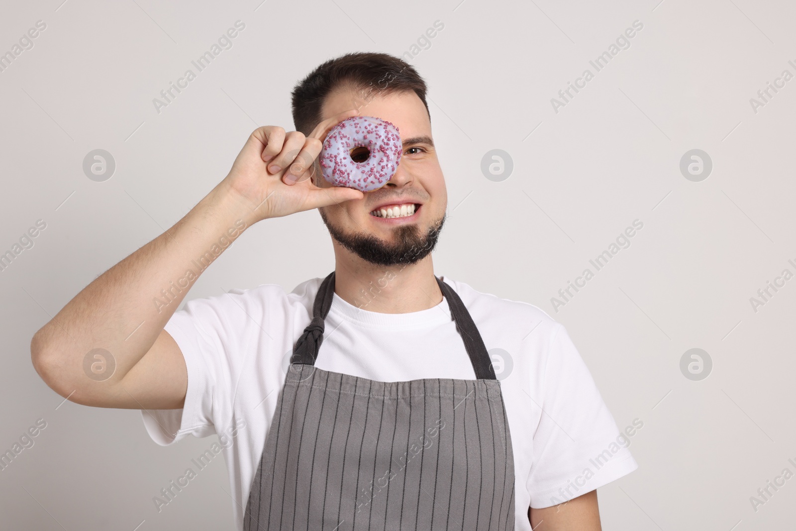 Photo of Happy professional confectioner in apron holding delicious doughnut on light grey background