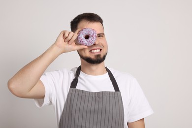 Photo of Happy professional confectioner in apron holding delicious doughnut on light grey background