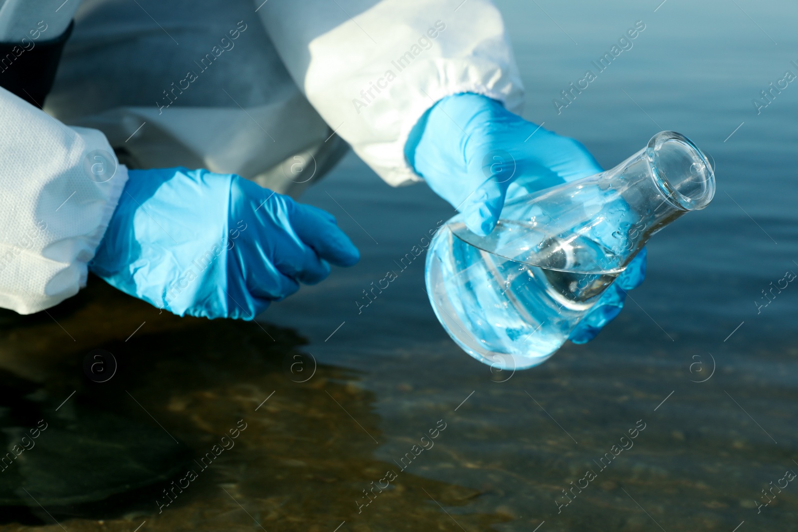 Photo of Scientist in chemical protective suit with conical flask taking sample from river for analysis, closeup