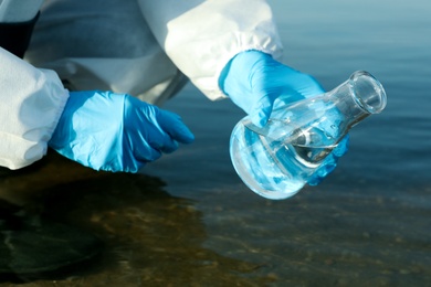 Scientist in chemical protective suit with conical flask taking sample from river for analysis, closeup