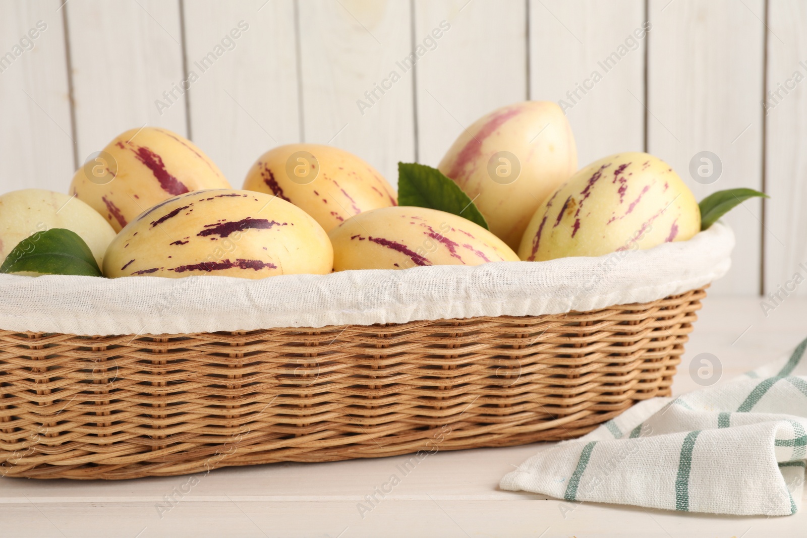 Photo of Fresh ripe pepino melons with green leaves in wicker basket on white wooden table