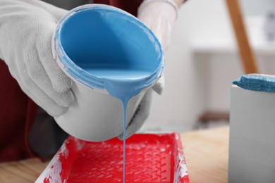 Photo of Woman pouring light blue paint from bucket into tray at table indoors, closeup. Space for text