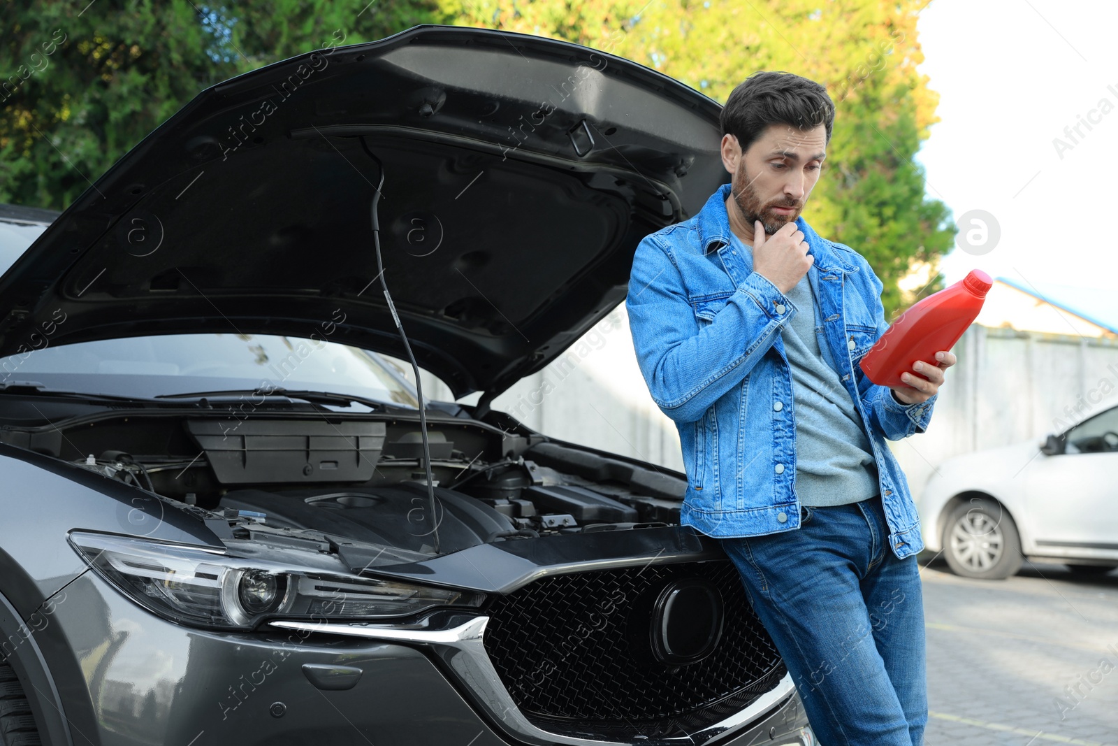 Photo of Puzzled man holding red container of motor oil near car outdoors