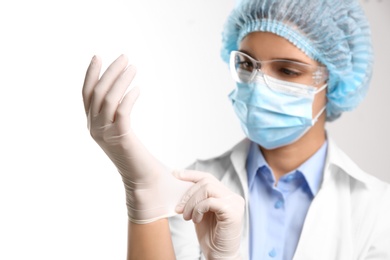 Woman in protective mask and glasses putting on medical glove against white background