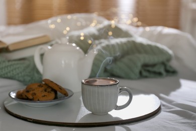Tray with cup of hot tea, cookies and teapot on bed indoors