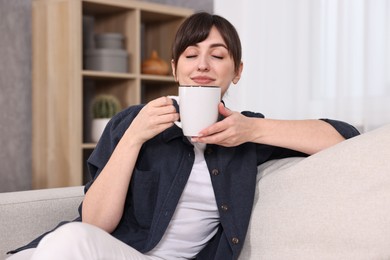 Beautiful young housewife with cup of drink on sofa at home