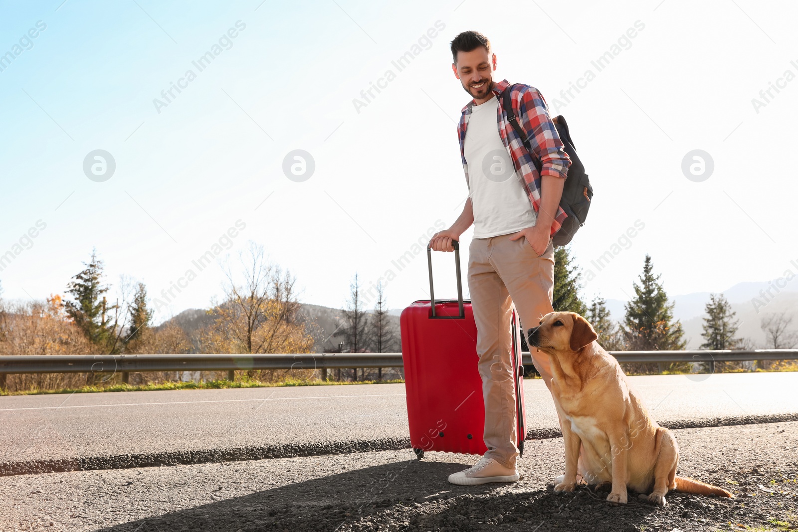 Photo of Happy man with red suitcase and adorable dog near road, space for text. Traveling with pet