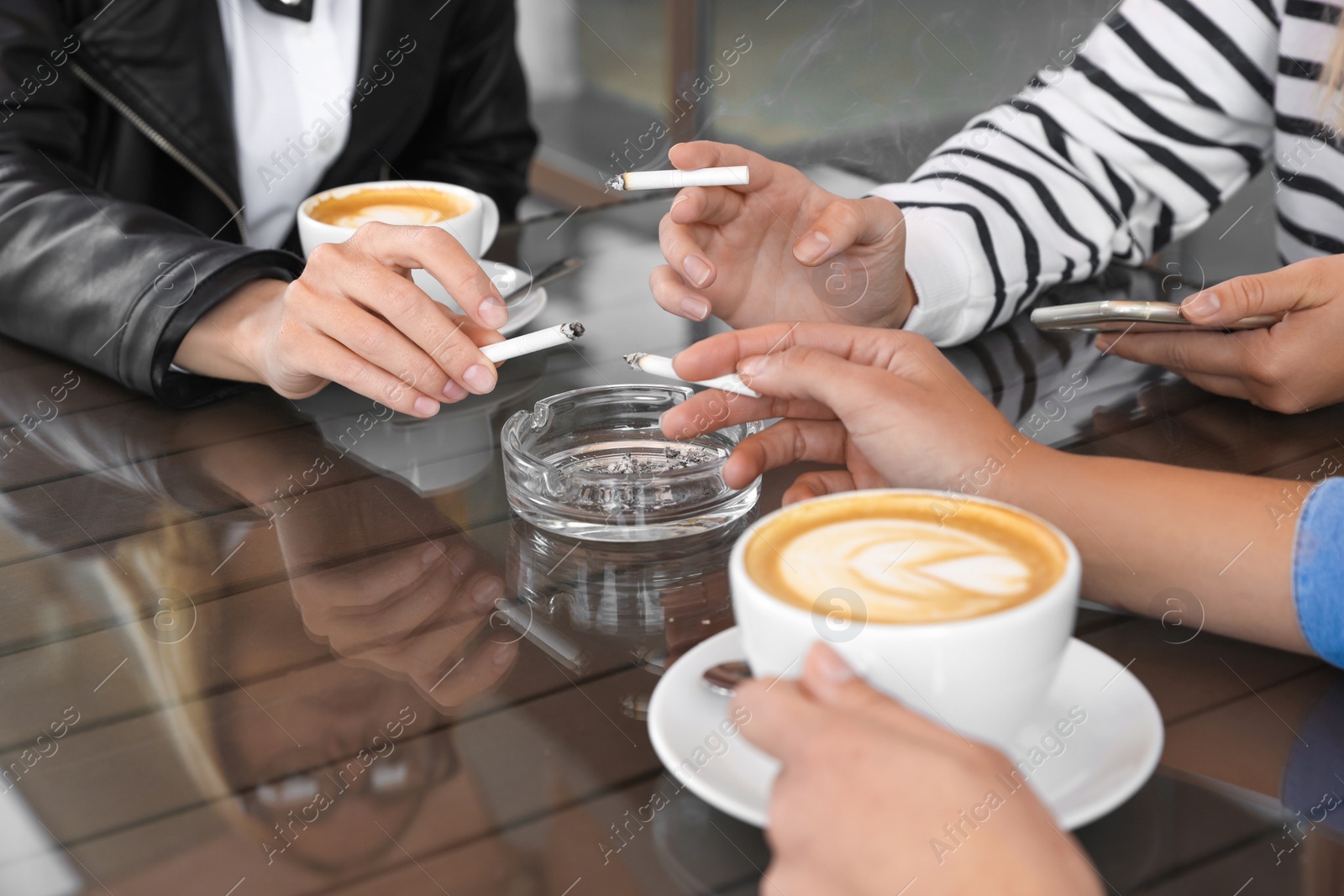 Photo of Women holding cigarettes over glass ashtray at table, closeup