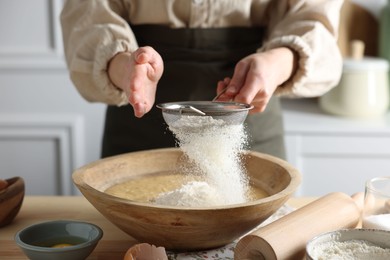 Making dough. Woman sifting flour into bowl at wooden table in kitchen, closeup