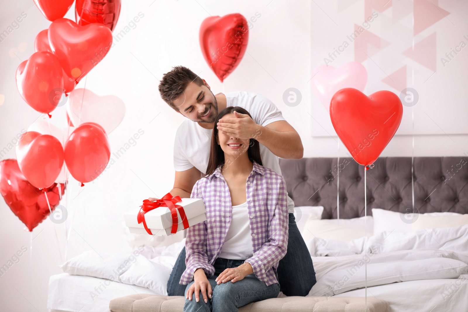 Photo of Young man presenting gift to his girlfriend in bedroom decorated with heart shaped balloons. Valentine's day celebration