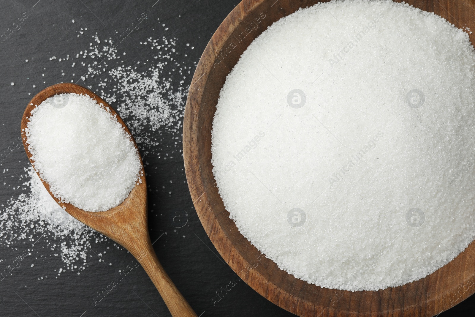 Photo of Granulated sugar in bowl and spoon on black table, flat lay