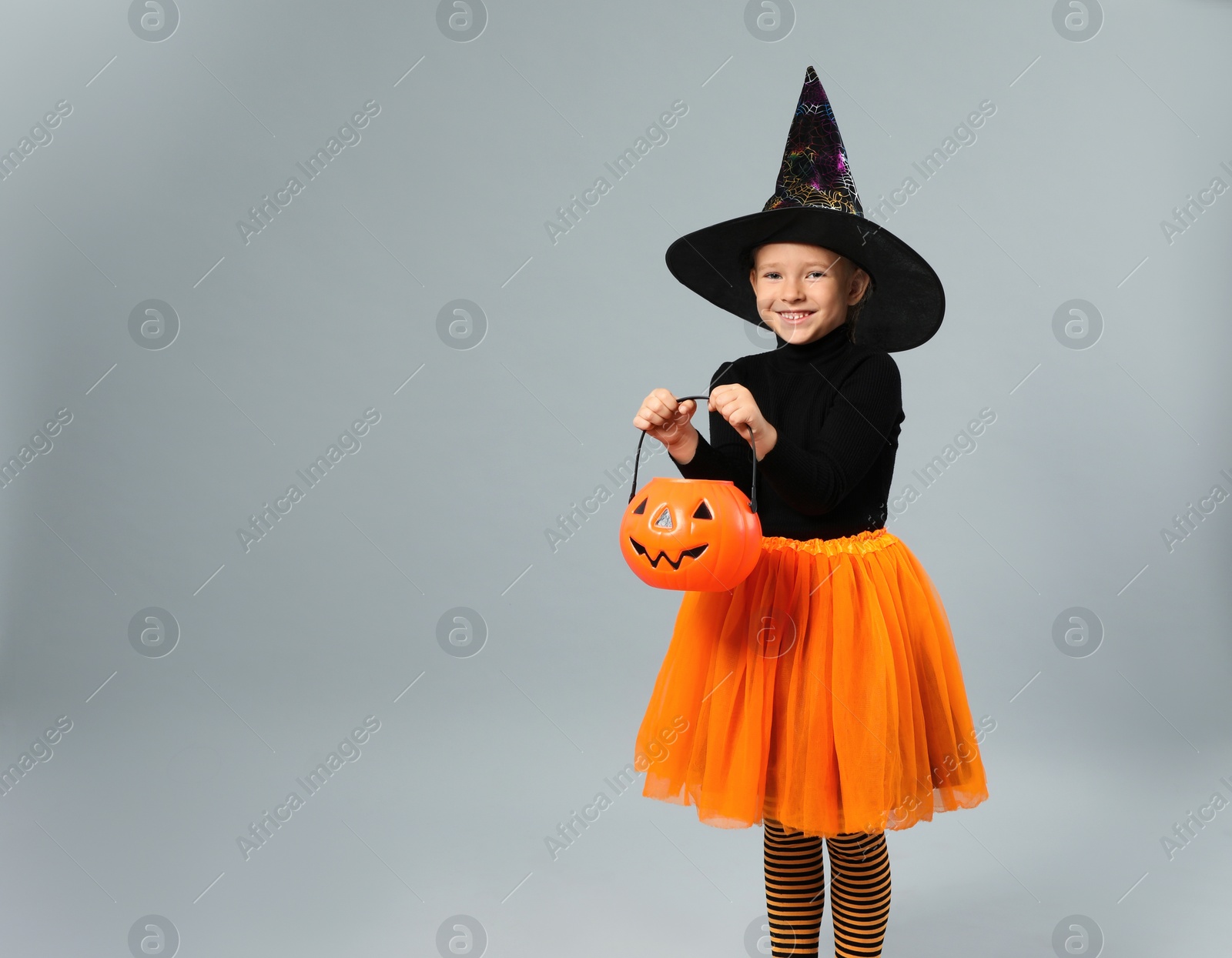 Photo of Cute little girl with pumpkin candy bucket wearing Halloween costume on grey background, space for text