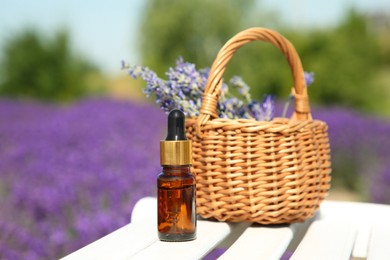 Bottle of essential oil and wicker bag with lavender flowers on white wooden surface outdoors, closeup