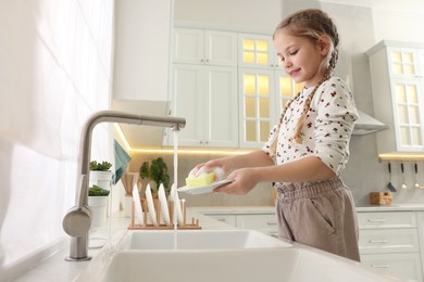 Little girl washing plate above sink in kitchen