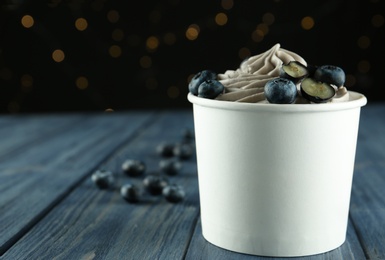 Photo of Cup of tasty frozen yogurt with blueberries on wooden table against blurred lights. Space for text