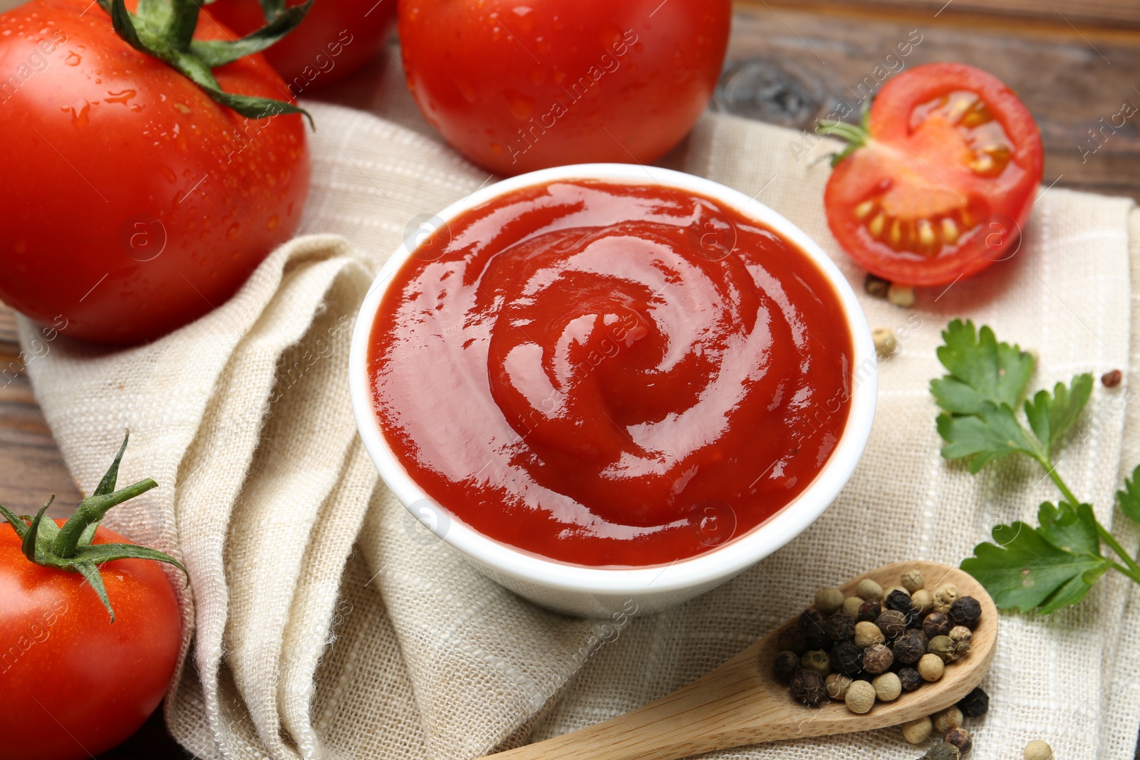 Photo of Delicious ketchup in bowl, tomatoes, parsley and peppercorns on table, closeup