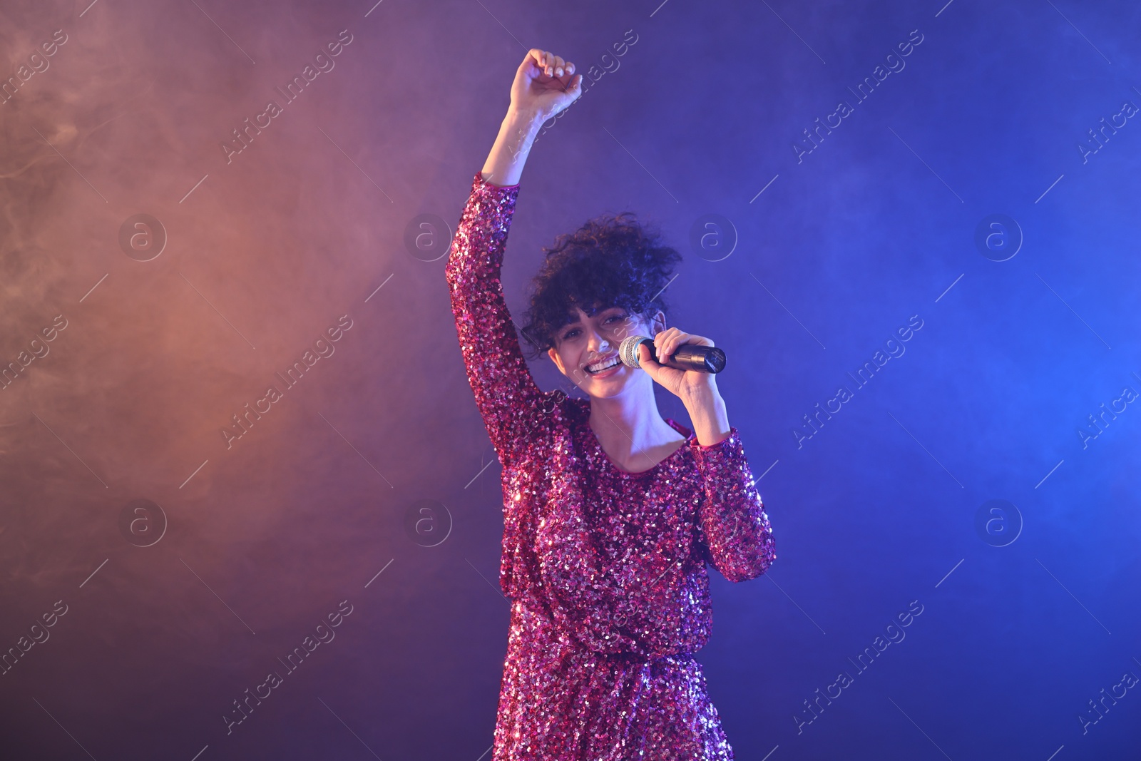 Photo of Beautiful young woman with microphone singing on color background in neon lights and smoke