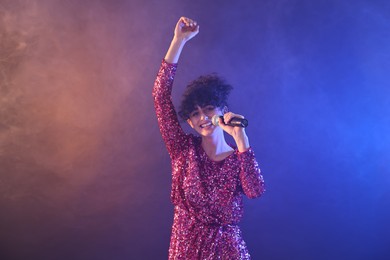 Beautiful young woman with microphone singing on color background in neon lights and smoke