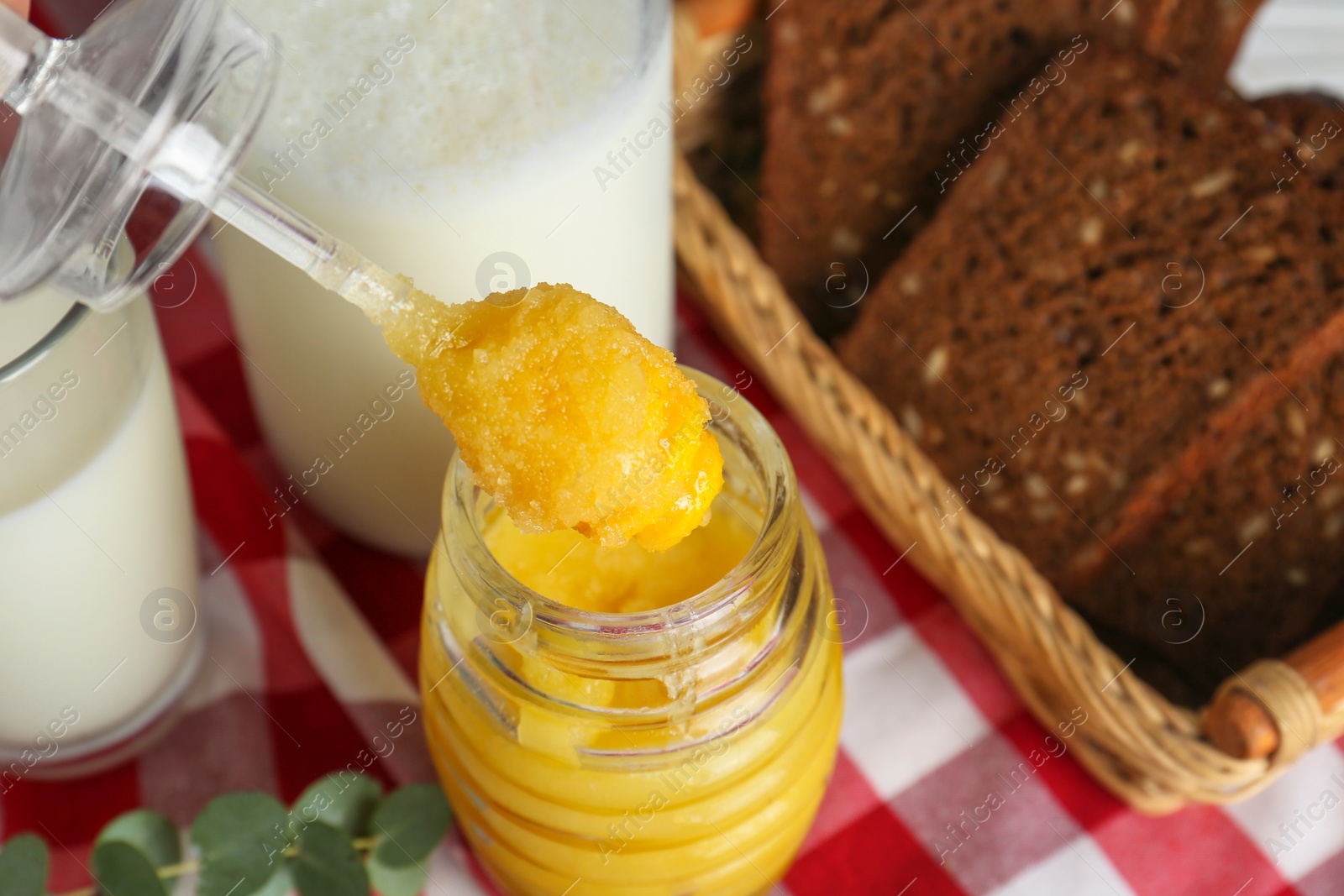 Photo of Taking tasty honey from jar with spoon near bread and milk on checkered cloth, closeup