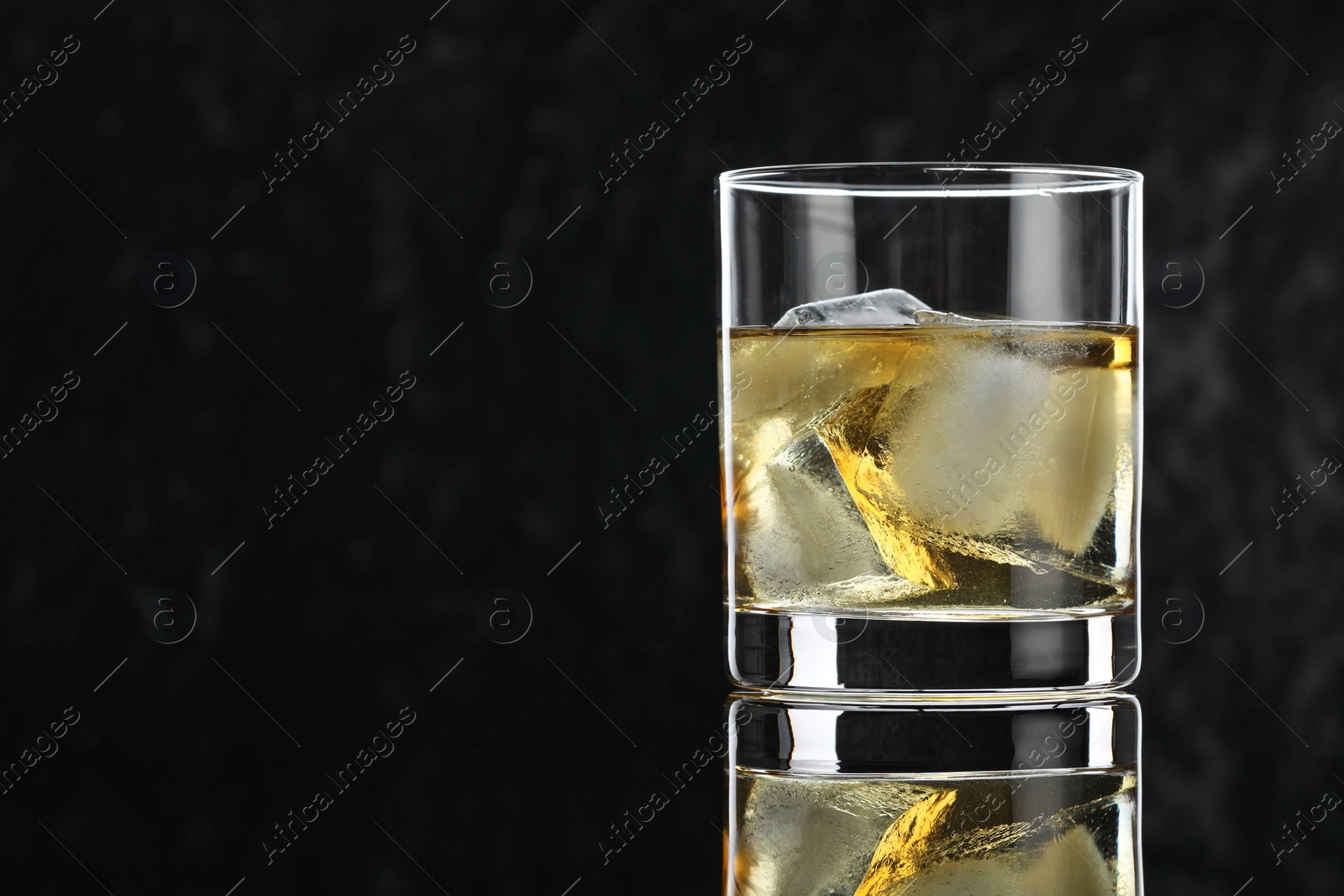 Photo of Tasty whiskey and ice cubes in glass on mirror table against black background, closeup. Space for text