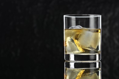 Photo of Tasty whiskey and ice cubes in glass on mirror table against black background, closeup. Space for text