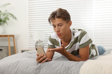 Photo of Happy young man having video chat via smartphone and blowing kiss on bed indoors. Long-distance relationship