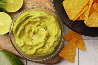 Bowl of delicious guacamole served with nachos chips and lime on white tiled table, flat lay