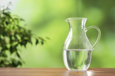 Photo of Glass jug with clear water on wooden table against blurred green background, closeup. Space for text