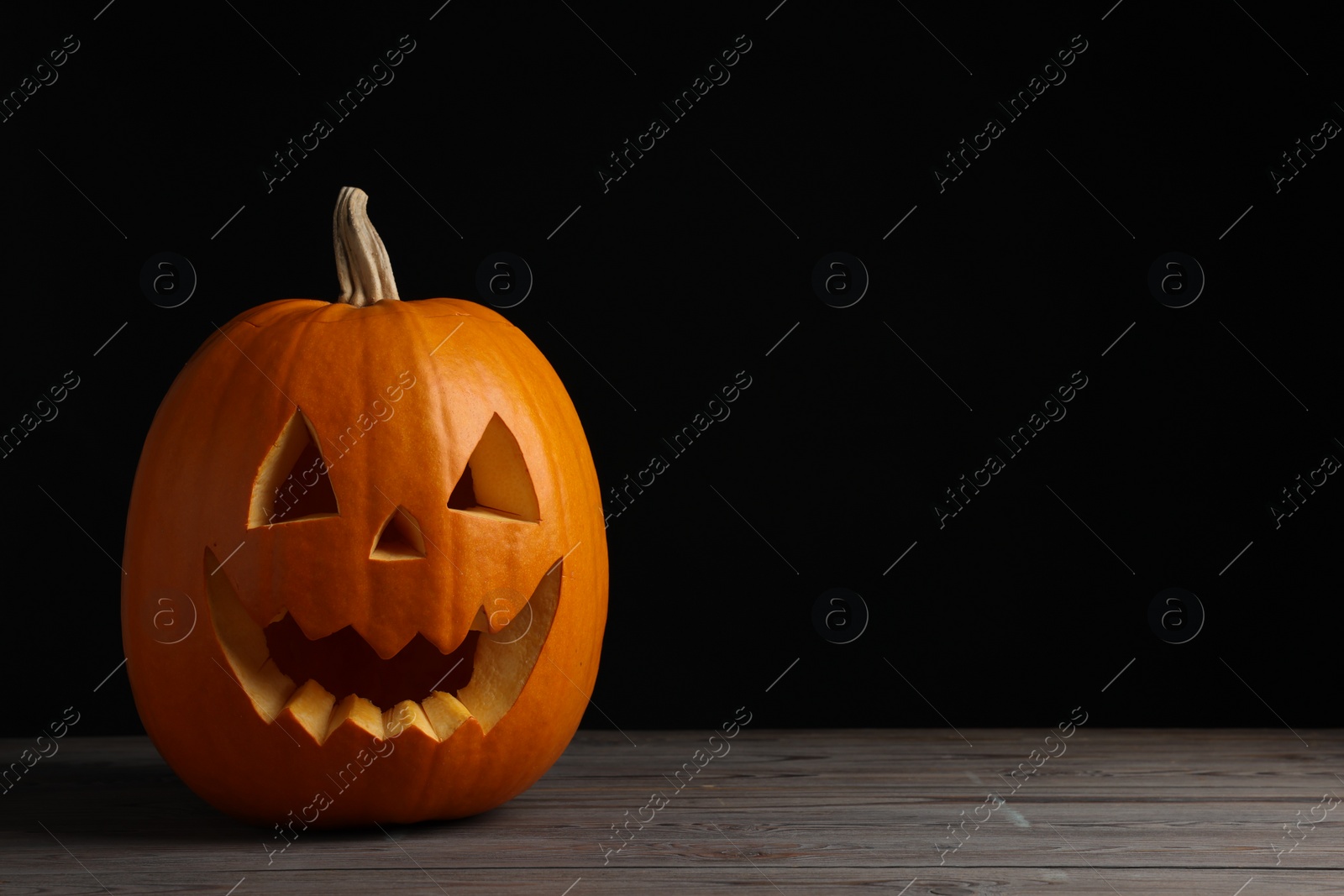 Photo of Scary jack o'lantern made of pumpkin on wooden table against black background, space for text. Halloween traditional decor