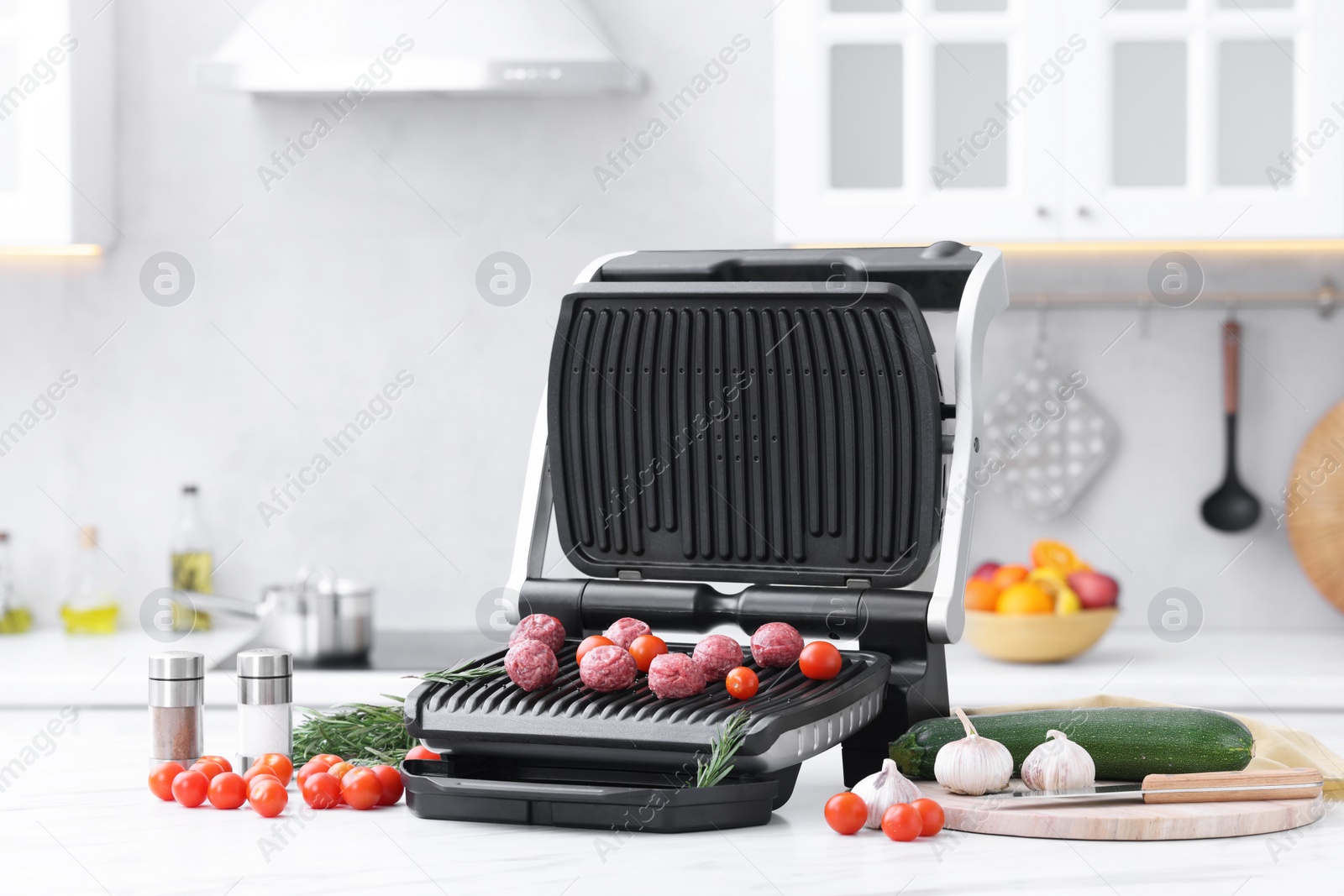 Photo of Electric grill with meatballs, tomatoes and vegetables on white marble table in kitchen