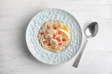 Photo of Plate of quinoa porridge with orange, banana and pomegranate seeds near spoon on white wooden background, top view