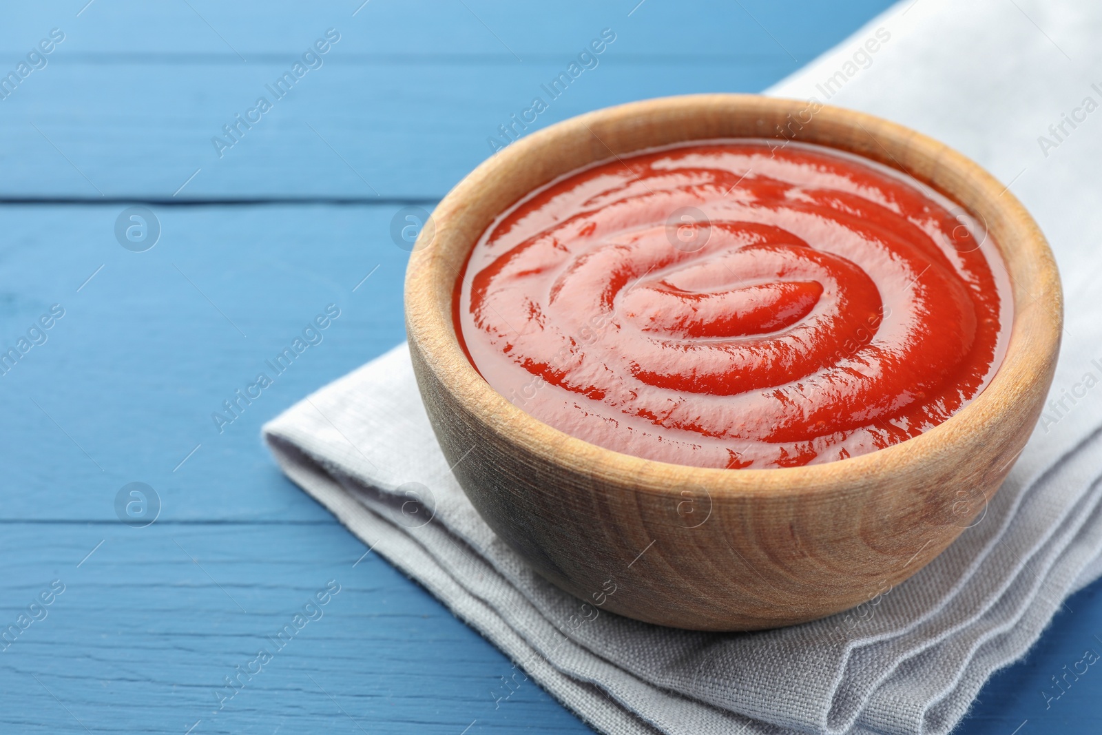 Photo of Bowl of tasty ketchup on blue wooden table, closeup. Space for text