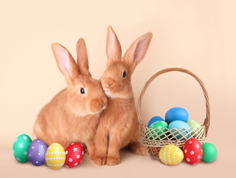 Cute bunnies and wicker basket with bright Easter eggs on beige background